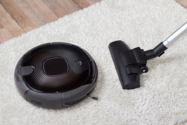 36316664 - traditional vacuum cleaner with a hose, nozzle and brush versus a modern circular automated low profile unit, high angle view on a shaggy white carpet